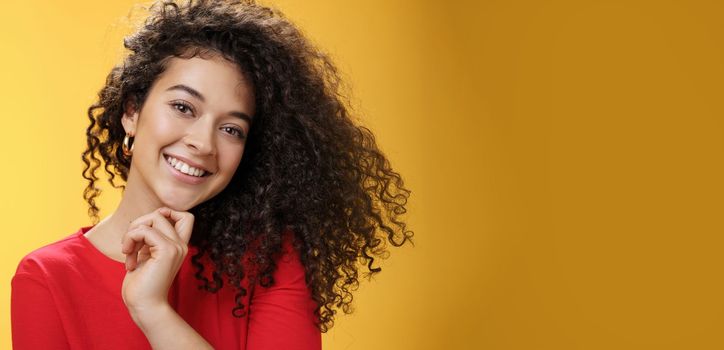 Lifestyle. Close-up shot of stylish and happy bright curly-haired female in red dress tilting head sensually touching chin with finger and smiling broadly making flirty gazed at camera over yellow background.