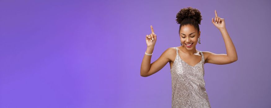 Good-looking carefree african-american girl student have fun prom dancing night club smiling delighted raicing index fingers up joyfully look down, triumphing celebrating good news, blue background.