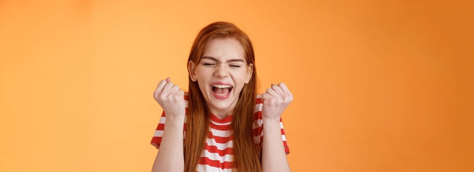 I did it. Charming lucky determined redhead female achieve success, fist pump celebrating, close eyes yell yes yeah, hooray gesture, triumphing victory, very good news, stand orange background.