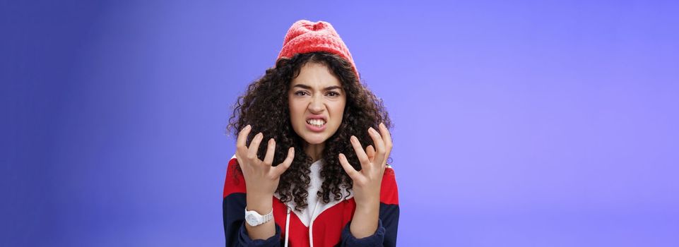 Pissed and annoyed stylish girl clenching hands in anger, grimacing squeezing teeth from annoyance and irritation, standing hateful and outraged feeling fury and hate posing over blue background.