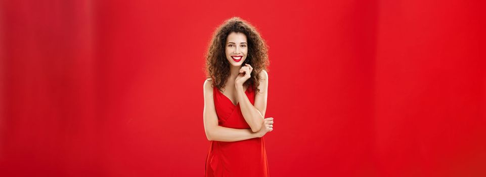 Passionate charming european woman over red background in elegant dress with curly hairstyle smiling cute, feminine playing with hair strand standing timid and silly, talking to person she admires.