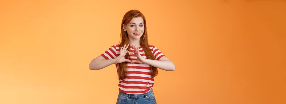 Cute lovely cheerful redhead girlfriend express love and cherish relationship, celebrating anniversary show heart sign, smiling tenderly, confess sympathy, stand orange background.