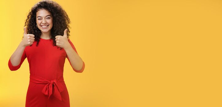 Creative and charismatic happy upbeat woman 25s with curly hair in red dress winking in approval and showing thumbs up with broad smile, satisfied giving positive reply over yellow wall.