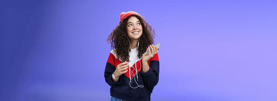 Lifestyle. Dreamy charming and happy kind girl recalling nice memories as listening nostalgic song in earphones holding smartphone looking at upper left corner with broad smile over blue background.