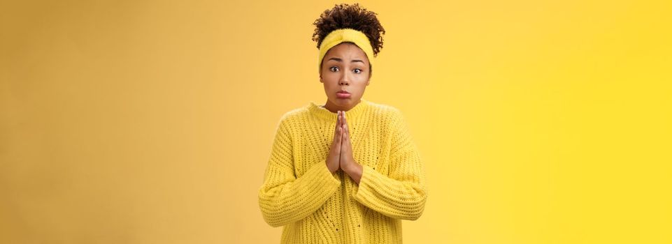 Silly cute pitty young african-american teenage daughter asking pocket money go trip press palms supplication pray need help begging make sad sulking miserable expression, standing yellow background.