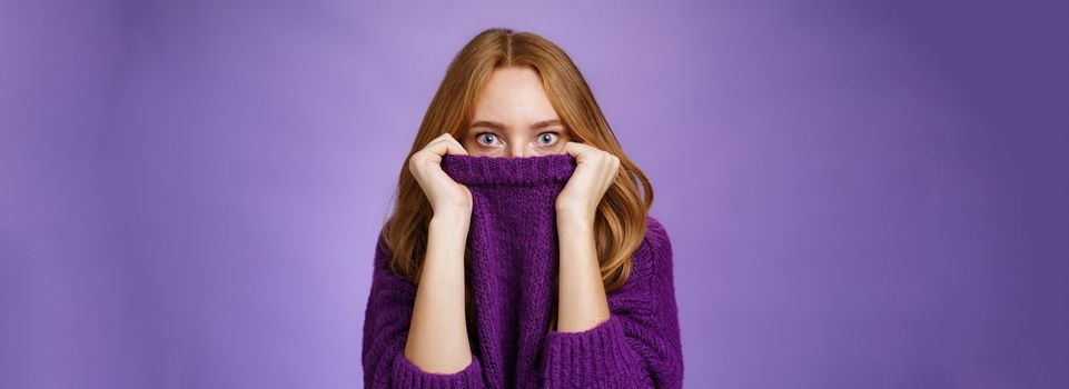 Lifestyle. Redhead girl scared winter is comming hiding in collar of warm sweater popping eyes shocked and stunned at camera as if frightened and insecure standing terrified over violet background.