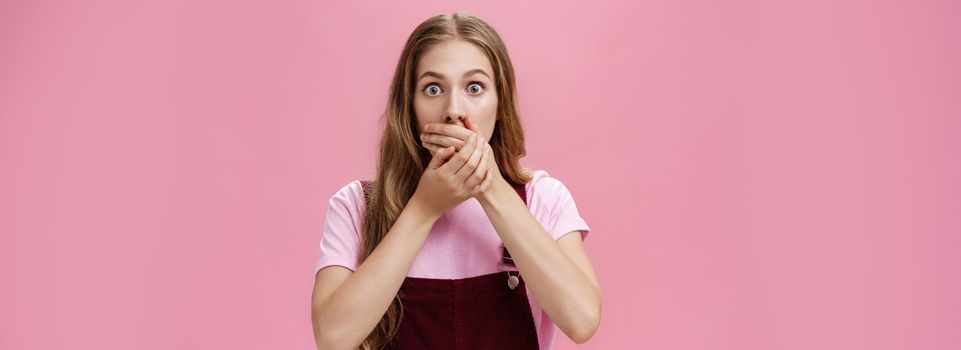 Lifestyle. Waist-up shot of shocked speechless pretty young girl in overalls with make-up covering mouth with palms from overreacting on terrible news witnessing accident nervously over pink background.