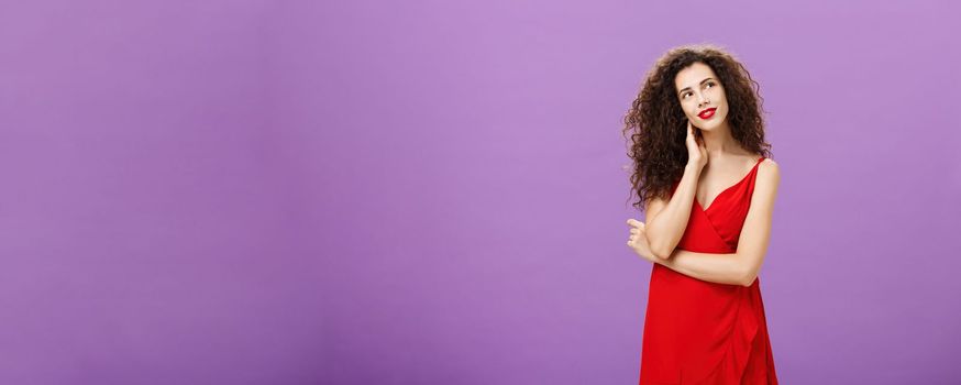 Charming and gentle elegant woman with curly hairstyle in stylish evening red dress tilting head touching neck and gazing at upper right corner tender, daydreaming posing over purple background.