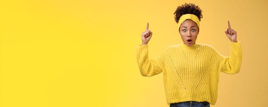 Shocked surprised emotive african-american female student drop jaw widen eyes learn astonishing news pointing raised fingers up gasping concerned impressed, speechless standing yellow background.