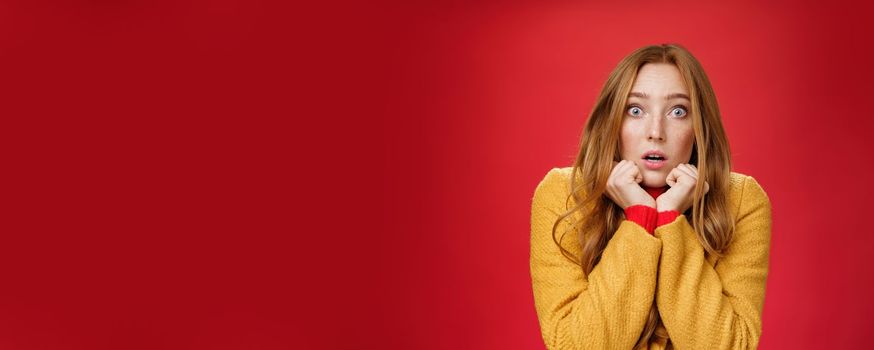 Close-up shot of shocked and scared insecure redhead woman in yellow coat pressing hands to jawline, open mouth and popping eyes at camera from amazement and fear over red background.