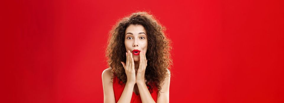 Charmed and intrigued curious good-looking rich woman in elegant dress with make-up and curly hairstyle folding lips in wow sign holding hands on face and smiling from excitement over red background. Emotions concept