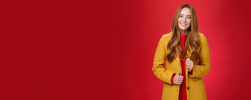 Stylish and cute good-looking redhead female in yellow coat on way to work, grabbing coffee making order with cute friendly smile touching button and posing over red background.