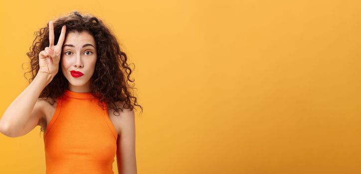 Waist-up shot of charismatic good-looking european female. with curly haircut in cropped top showing peace sign near face making silly look smirking and gazing friendly at camera over orange background.