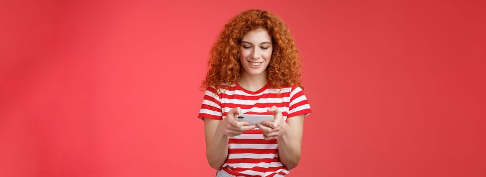 Lifestyle. Redhead girl fool around waiting queue dentist playing awesome smartphone game hold phone horizontal tap cellphone screen look telephone display smiling delighted entertained standing red background.