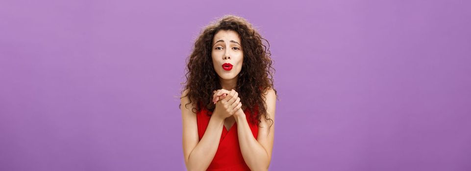 Waist-up shot of woman. gazing at heartwarming scene being touched and delighted holding palms togehter over chest folding lips enjoying watching cute puppy posing pleased over purple background.