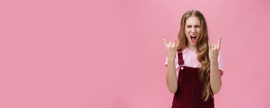 Cute and excited party girl with wavy hairstyle in corduroy overalls yelling yeah and showing rock-n-roll gesture having fun enjoying hard music being daring and rebellious over pink wall. Copy space