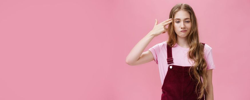 Portrait of bothered uneasy cute european female with long natural fair hair in trendy corduroy overalls smirking from irritation and annoyance showing finger fun gesture as if shooting in head. Copy space