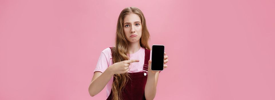 Lifestyle. Sad gloomy young female with cute wavy natural hairstyle showing smartphone screen pointing at gadget with index finger making upset face frowning displeased feeling regret after buying broken phone.
