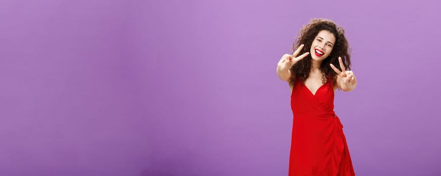 Friendly-looking peaceful european female with curly haircut in elegant red dress showing peace or victory signs, smiling broadly saying her enjoying awesome party in friends circle over purple wall. Copy space