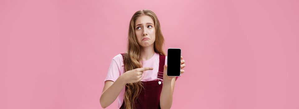 Indoor shot of gloomy displeased and disappointed cute young girl holding smartphone pointing at cellphone screen making upset grimace as if being bothered and dissatisfied against pink wall. Emotions and technology concept