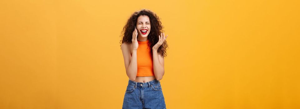 Sociable stylish and popular european woman with curly hairstyle and red lipstick laughing out loud having fun talking on smartphone with friend playing with hair strand carefree over orange wall. Technology concept