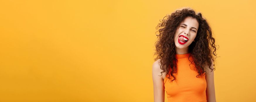 Portrait of upbeat playful and stylish caucasian girl. with curly hair and red lipstick winking showing tongue with rebellious hot expression tilting head standing cool and chill over orange background.