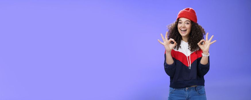 Got under control. Portrait of happy charming smiling curly-haired female in warm winter hat and sweatshirt smiling broadly and showing okay or excellent gesture as approving, liking cool movements.