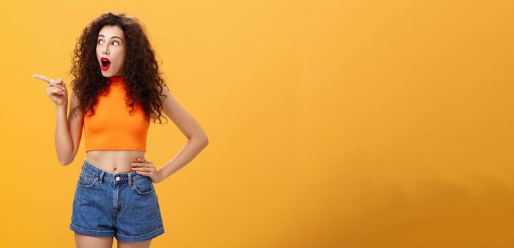 Woman peeking at neighbours window. being surprised and amazed pointing and staring speechless left standing over orange background with dropped jaw intrigued and thrilled look in cropped top.