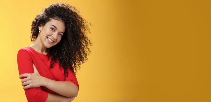 Waist-up shot of tender feminine and gentle woman with curly hairstyle combed to right side, tilting head and smiling flirty making romantic gazed at camera hugging herself over yellow background.