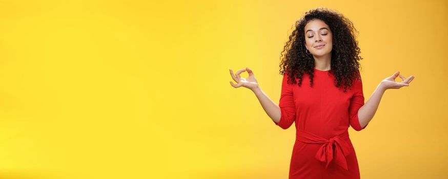 Charming dreamy and happy peaceful woman in red dress with curly hair standing in lotus pose with mudra orbs or zen gesture and closed eyes, meditation relieving stress, feeling unbothered. Body language, buddhism and people concept