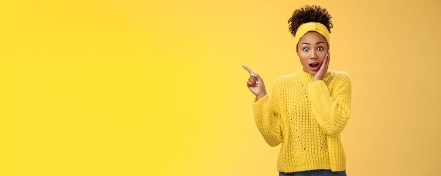 Shocked impressed emotive african-american girl afro hairstyle. gasping amazed touch cheek stunned widen eyes pointing upper left corner surprised telling you unbelievable discovery yellow background.