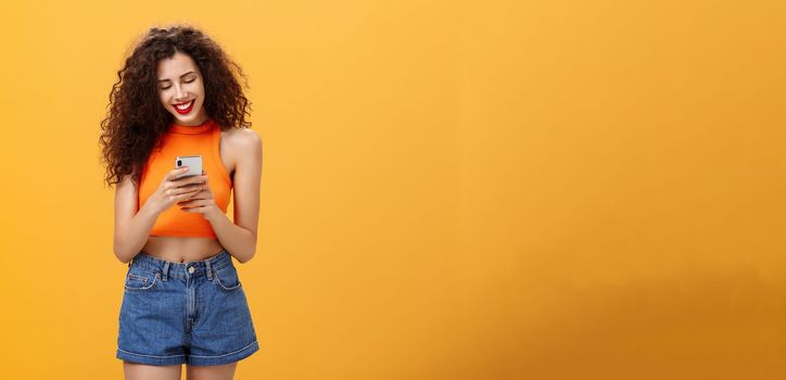 Girl spending time in internet texting friend messages. via smartphone laughing while looking at device screen standing happy and upbeat over orange background in cropped top and denim shorts.