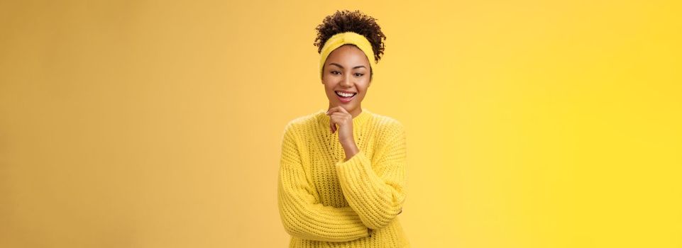 Interested excited enthusiastic good-looking female entrepreneur stylish sweater headband afro hairstyle touching chin thoughtful smiling liking project idea standing yellow background intrigued.