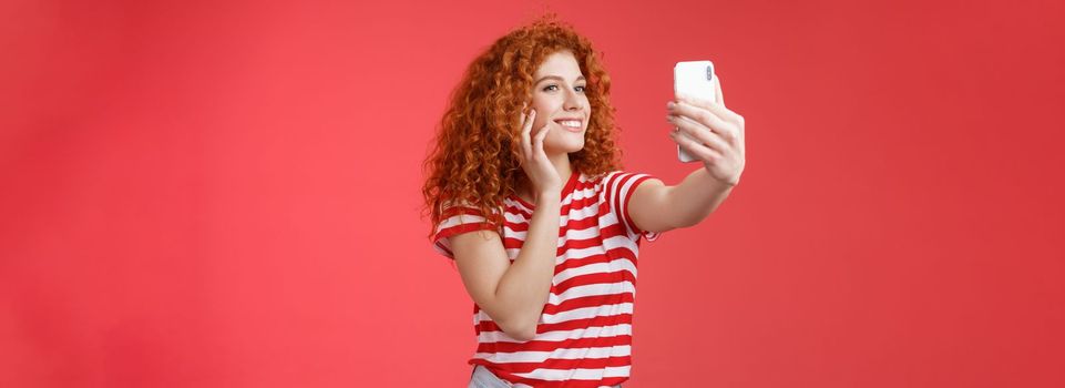 Lifestyle. Popular cheerful good-looking stylish female blogger redhead curly hairstyle feel pretty self-acceptance taking selfie raised arm holding smartphone posing silly cute phone camera red background.