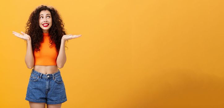 Silly and clueless carefree cute girl. with curly hairstyle in cropped top and shorts shrugging with palms spread aside smiling unaware and gazing at upper right corner uncertain and uninvolved.