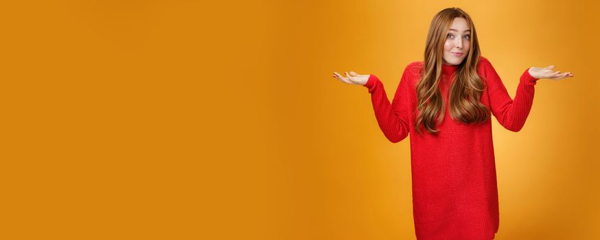 What do I know. Portrait of confused clueless tender and gentle redhead woman in red sweater shrugging with hands raised near shoulders turning away questioned and unaware, not knowing.