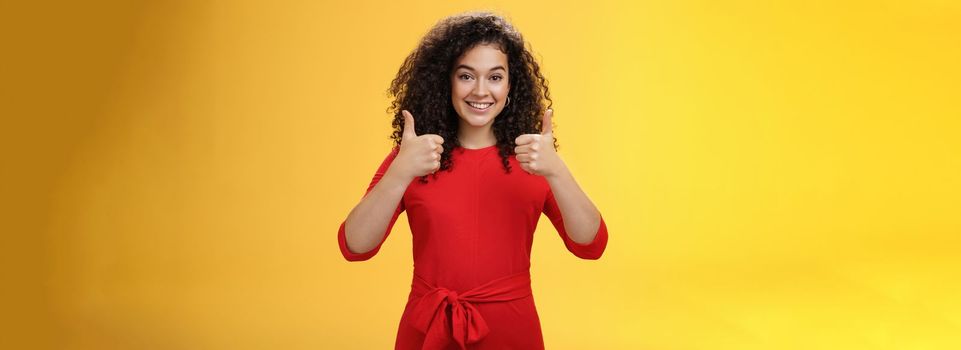 Excellent idea, nice job. Portrait of supportive delighted and happy charming female friend showing thumbs up as standing in red dress over yellow wall smiling giving positive reply, liking concept.