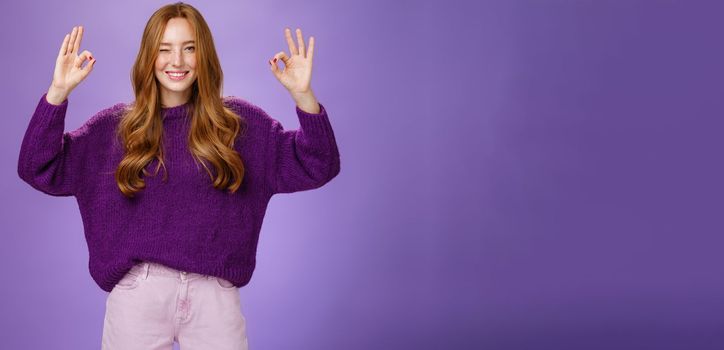 Relax everything ok. Assured charming and happy young redhead with long beautiful hair and freckles winking joyfully smiling and showing okay gestures with raised hands, affirming everything perfect.