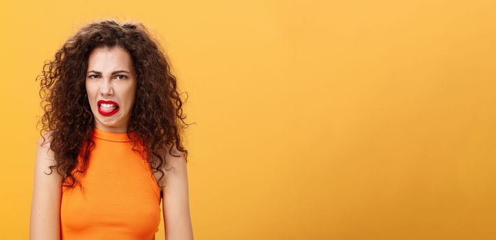 Girl being disgusted with row food sticking out tongue and frowning showing aversion and disgust talking about thing she dislikes with friend standing intense and dissatisfied over orange background. Copy space