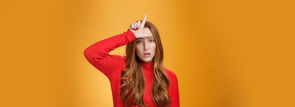 Arrogant and self-satisfied redhead woman humiliating person showing loser sign on forehead mocking and disdain rival standing confident and snobbish over orange background. Copy space