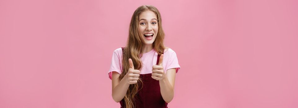 Friendly and outgoing pretty young woman in overalls with wavy natural hairstyle showing thumbs up gesture with cheer and joy smiling being supportive, liking idea, expressing positive opinion. Gestures concept