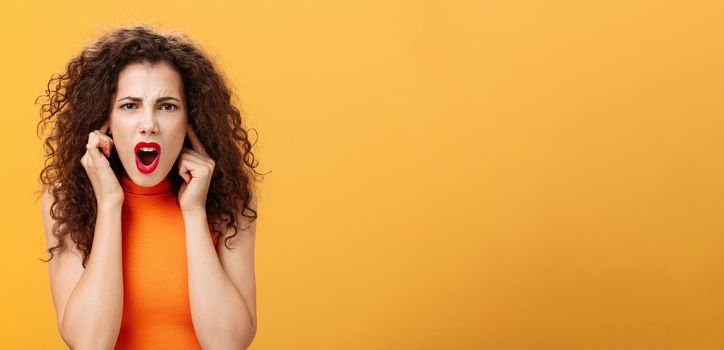 Waist-up shot of displeased intense and unsure female. with curly hairstyle in orange top closing ears with fingers frowning asking repeat question while standing in loud noisy place over orange wall.