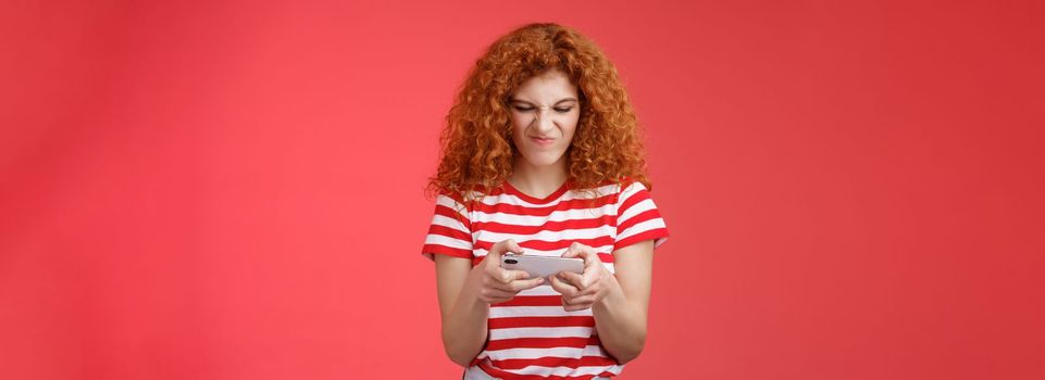 Daring enthusiastic addicted geeky good-looking redhead curly girl hate losing battles smartphone game wrinkle nose grimacing upset eager win tap fast telephone screen look display, red background.