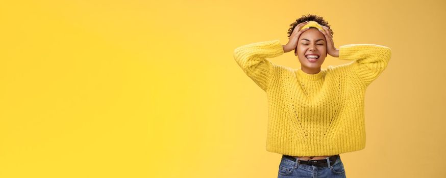 Carefree happy lucky african girl in sweater headband touching head having fun smiling relieved excited laughing out loud close eyes rejoicing spend good time partying, standing yellow background.