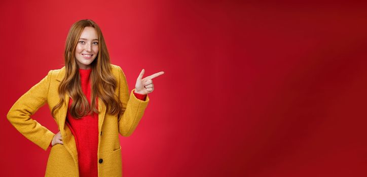 Lifestyle. Stylish and self-assured attractive redhead woman with freckles in yellow outdoor coat pointing right with finger pistol and smiling joyfully as camera as showing cool promotion over red wall.