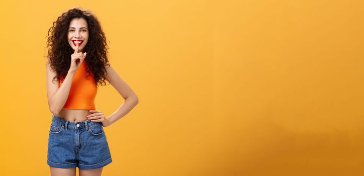 Shh I got something for you. Portrait of charming european young female with curly hairstyle in cropped top and shorts showing shush gesture with broad smile having secret preparing surprise.