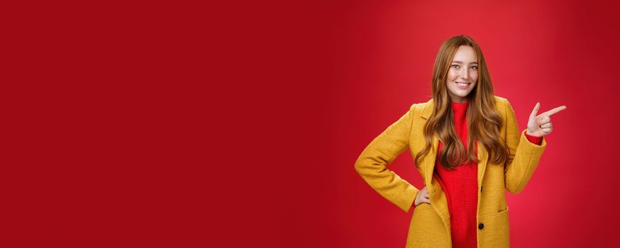 Lifestyle. Stylish and self-assured attractive redhead woman with freckles in yellow outdoor coat pointing right with finger pistol and smiling joyfully as camera as showing cool promotion over red wall.