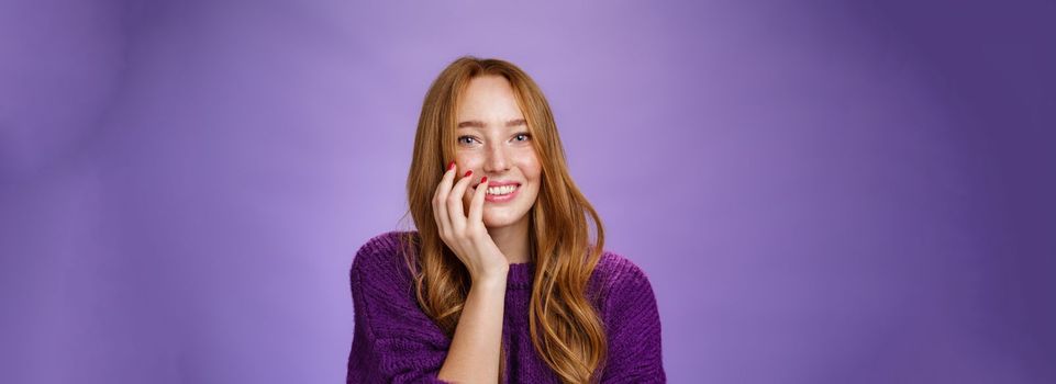 Lifestyle. Close-up shot of feminine and glamour cute redhead girl with freckles in purple knitted sweater holding hand on cheek gently chuckling and smiling broadly at camera over violet background.