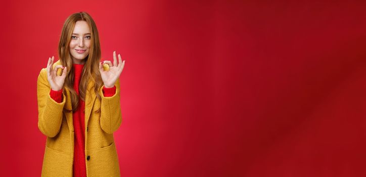 Indoor shot of pleased bright and happy young woman feeling okay and perfect in warm comfortable yellow coat picking right close for cold weather showing ok gesture and smiling delighted over red wall. Fashion and temprature concept