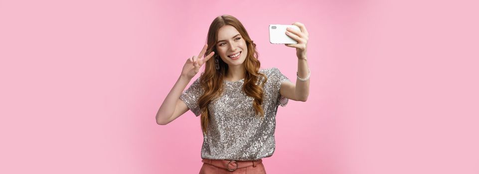Sociable attractive glamour young caucasian woman taking selfie show victory peace sign smiling broadly raise smartphone look grinning mobile phone display, video calling friends, pink background.